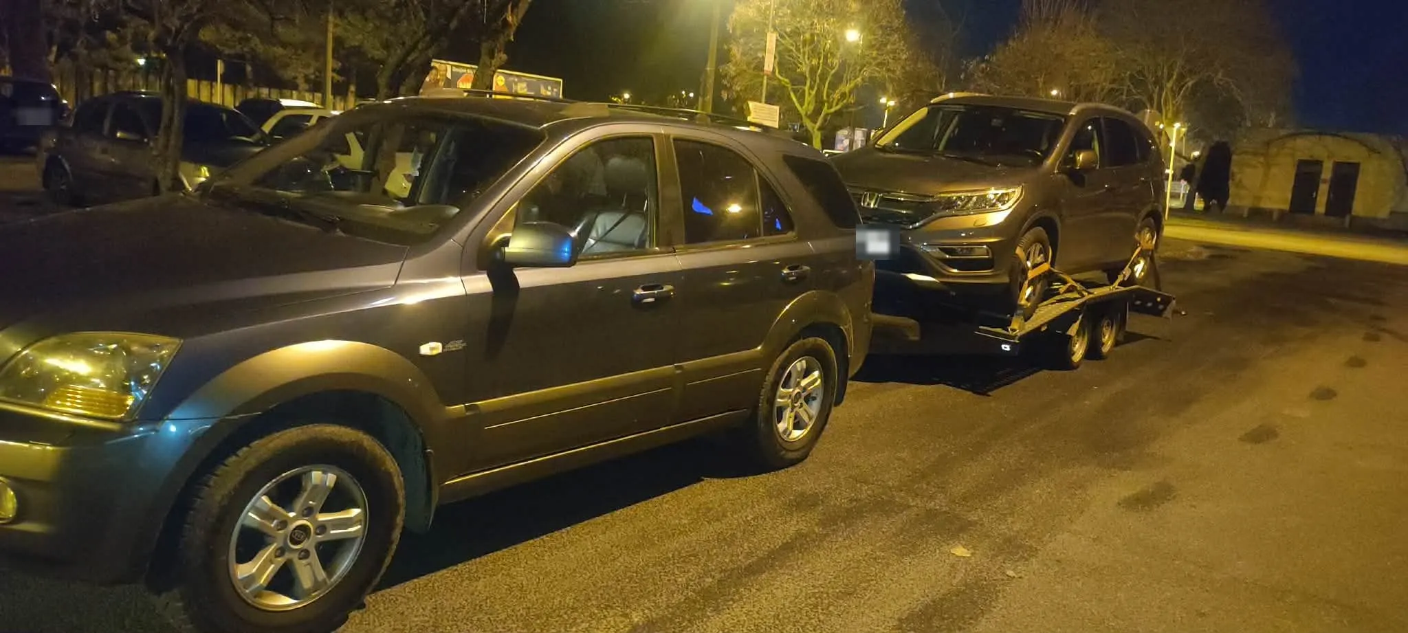 A vontató terepjáró éjszaka, mögötte az autószállító trélerre rögzített szürke SUV