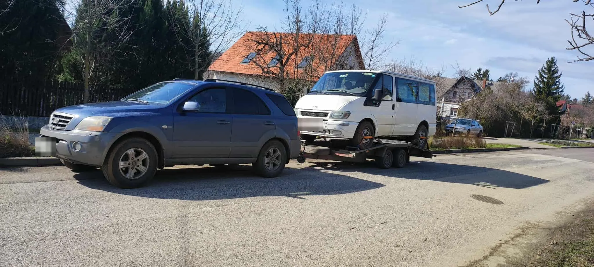 Fehér kisbusz a trélerre rögzítve, a vontató terepjáró mögött