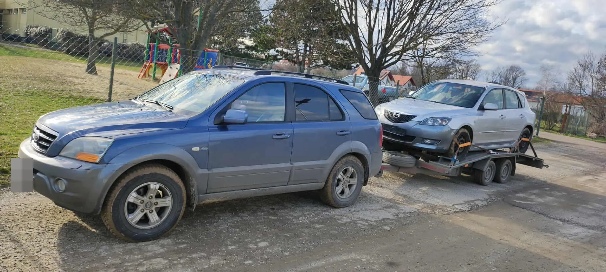 Ezüst színű, üzemképtelen személyautó a trélerre rögzítve, a vontató terepjáró mögött